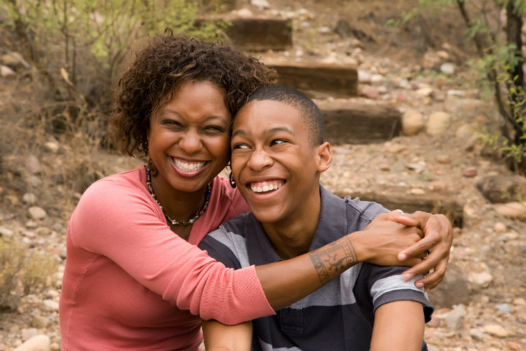 Image of a Black woman in a red shirt hugging a Black teenaged son in a gray shirt. They are standing in front of outdoor stairs.