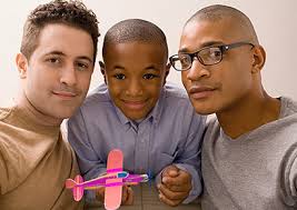 Image of a family. On the left is a man with dark hair and a brown shirt. in the center is a Black child in a blue button up shirt, holding a pink and purple toy plane. On the left is a Black man in a gray shirt and glasses. 
